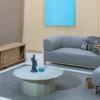 A modern living room features a gray sofa, matching ottoman, Timber TV stand, and a Karimoku New Standard, Colour Wood Coffee Table with decorative items. Walls are light wood with a teal abstract painting. Rug covers part of the floor.